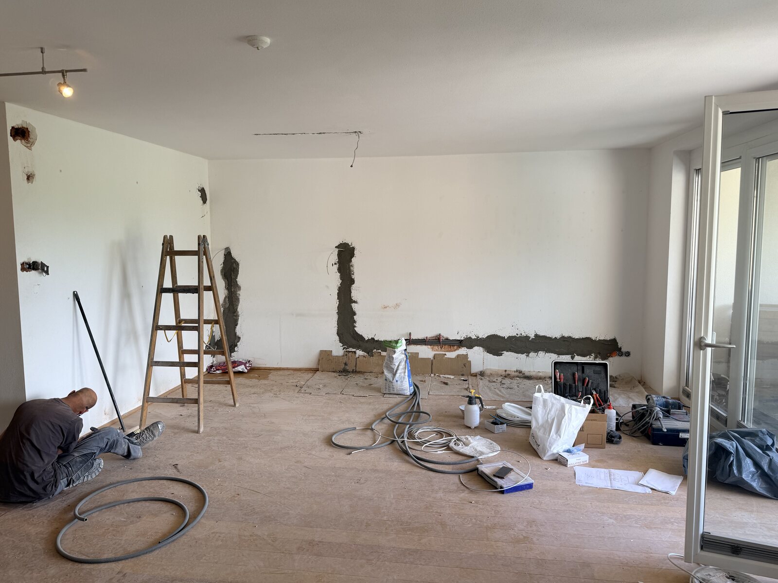 Wohnung in Sanierung: frisch verschlossene Wandschlitze, Leiter, Werkzeug und Kabel auf dem Rohboden, links arbeitet jemand an der Wand
